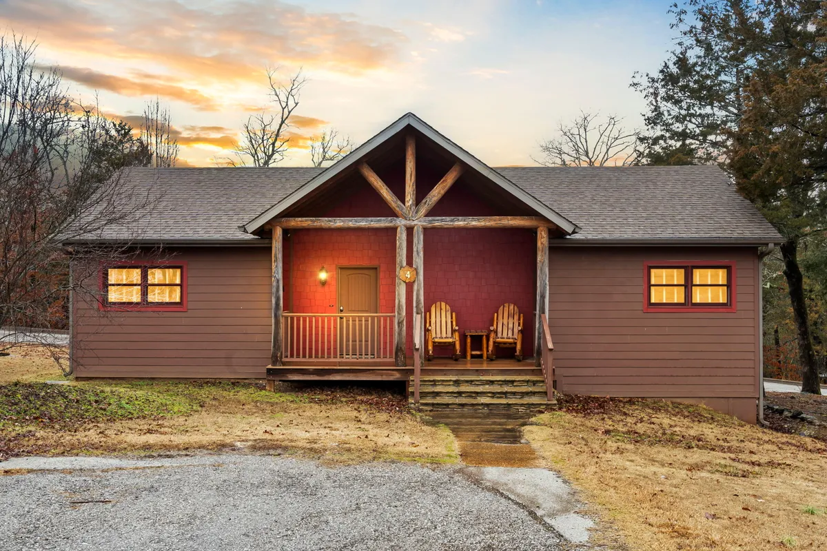 A spacious wooden porch with welcoming rocking chairs, red accents framing the front door, and lush landscaping set the scene. The area is designed for gathering, with ample space for friends to relax, sip drinks, or enjoy sunset views. The porch exudes a cozy, communal atmosphere perfect for shared al fresco dinners or evening conversations.