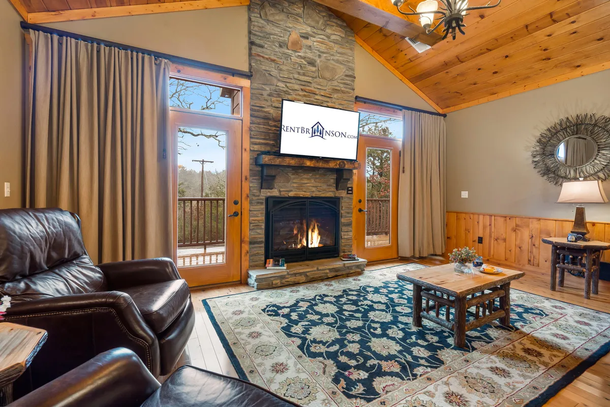 A cozy living room with a glowing propane fireplace, a soft area rug, exposed wooden beams, and large windows that look out onto nature. The seating area invites relaxation and connection.