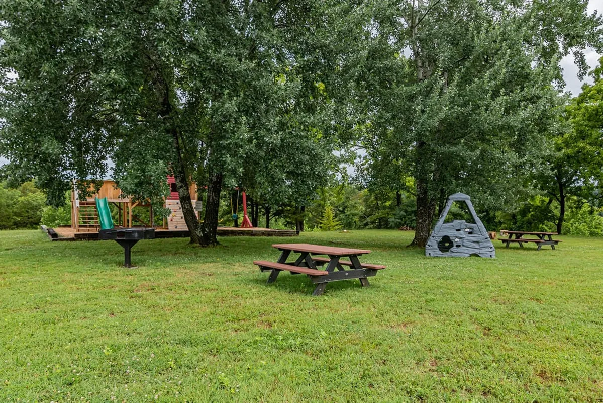 This image showcases a vibrant outdoor playground complete with a play structure, swings, climbing wall, picnic tables, and a grill set among trees. Children can be seen enjoying the play area while families make use of the picnic facilities, suggesting a lively and welcoming atmosphere perfect for families seeking outdoor fun and togetherness.