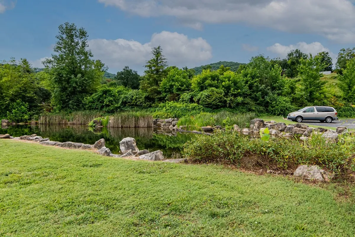 A serene waterscape with a reflective water surface, lush greenery, rock formations, and a parked vehicle off to the side. The scene exudes peacefulness and the beauty of untouched nature, suggesting opportunities for kayaking, fishing, and wildlife observation along the water’s edge.