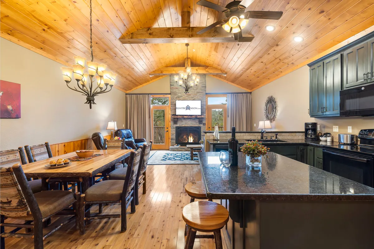 The image features a cozy, adirondack-style interior with a glowing fireplace, a rustic wooden dining table set for family or friends, a beautifully lit chandelier, and an open kitchen complete with a spacious island. The warm ambiance invites togetherness and relaxation, perfectly matching the lodge's peaceful lakeside setting.
