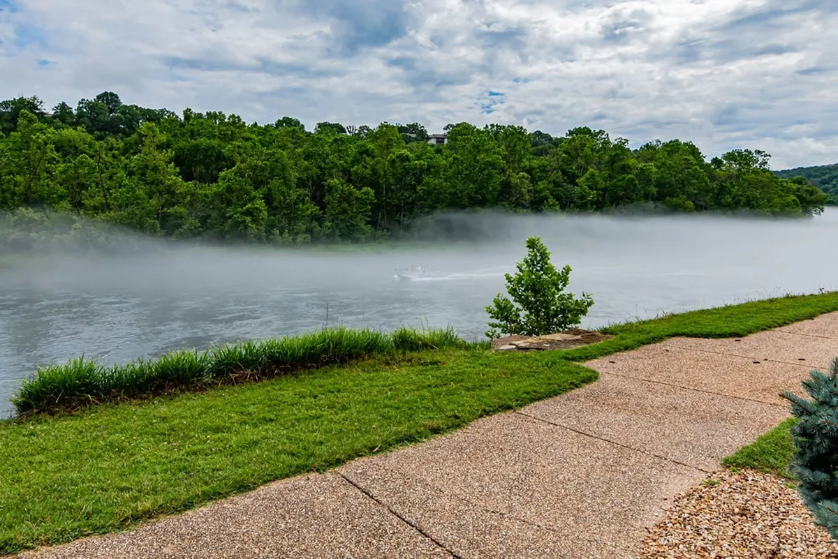 A peaceful riverside scene with a misty river, lush green trees, and a walking path alongside the water. The natural environment looks serene, ideal for quiet strolls and peaceful reflection, and it conveys the proximity to Lake Taneycomo and outdoor activities.
