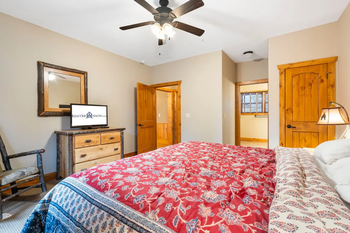 A welcoming bedroom highlighted by a king bed with red patterned bedding, a large wooden dresser, mirror, and a cozy rocking chair by the window. The space feels warm and homely, with a television for entertainment and a glimpse of natural light.