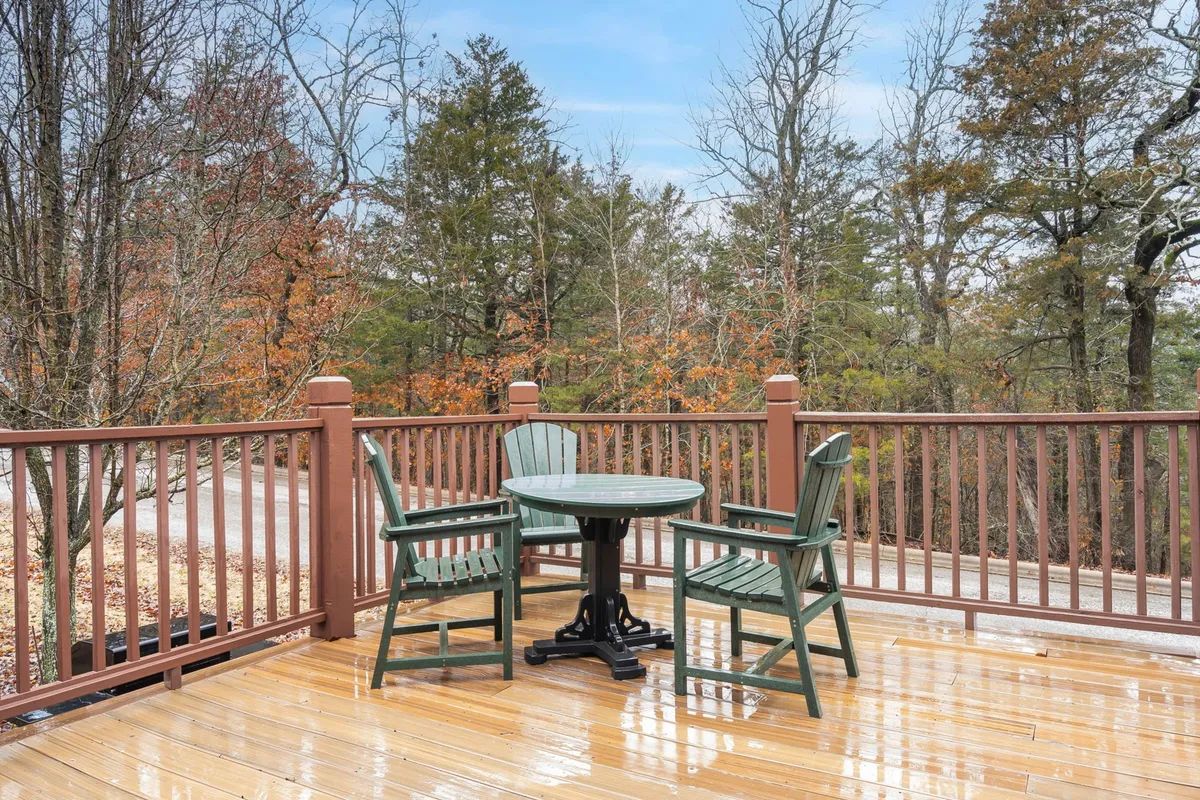 A wooden deck with green chairs and a round table sits nestled among the trees. The setup is ideal for enjoying coffee in the morning, reading, gathering with friends, or dining al fresco.
