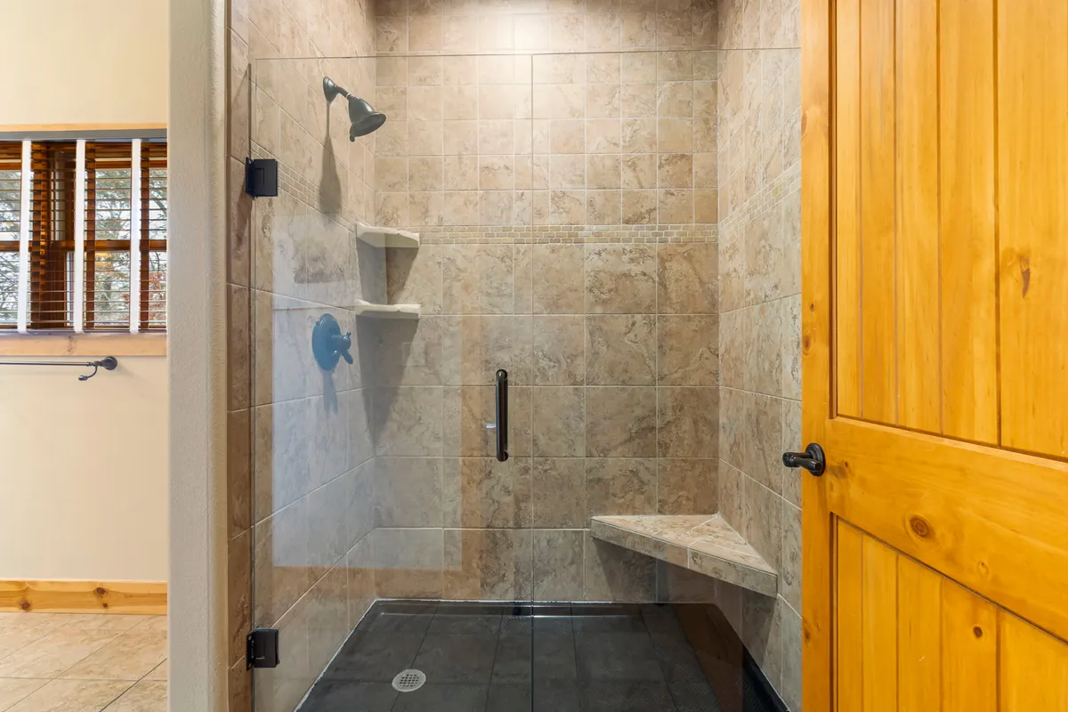A modern bathroom with a spacious glass shower enclosure, natural stone tiles, and a wooden door. The area exudes a tranquil, spa-like atmosphere, ideal for unwinding after a day of adventure.