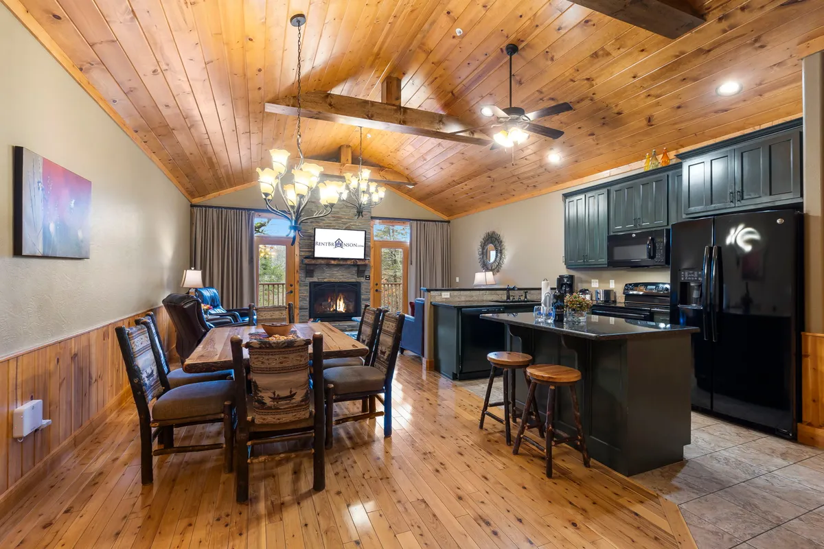 This image features a welcoming open-concept space that highlights the dining table, a cozy fireplace, the kitchen island, and a chandelier overhead. The mix of wood tones and soft lighting creates an inviting atmosphere, and the layout clearly connects the kitchen, dining, and living areas.